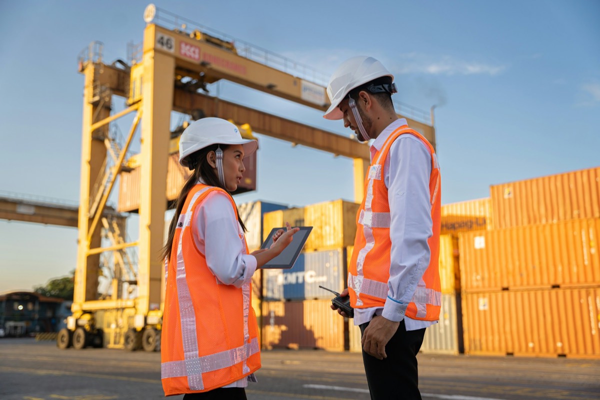 Two engineers in hard hats checking a tablet beside shipping containers and a gantry crane